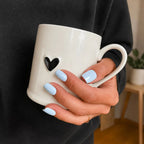 Manucure Press-on Bleu Perle carrés courts sur main tenant un mug blanc coeur noir, sweat gris et plante verte en fond, bague fine dorée, ongles bleu ciel doux et cosy LYLY Nails