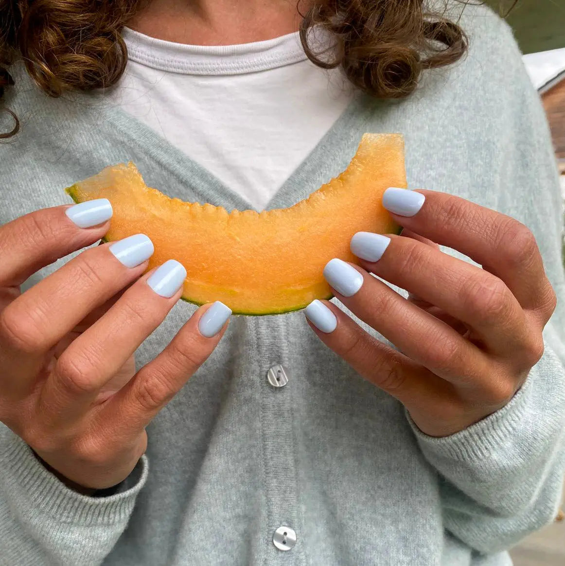 Faux ongles Bleu Perle carrés courts sur deux mains tenant une tranche de melon, gilet gris clair et t-shirt blanc, ambiance estivale fraîche LYLY Nails