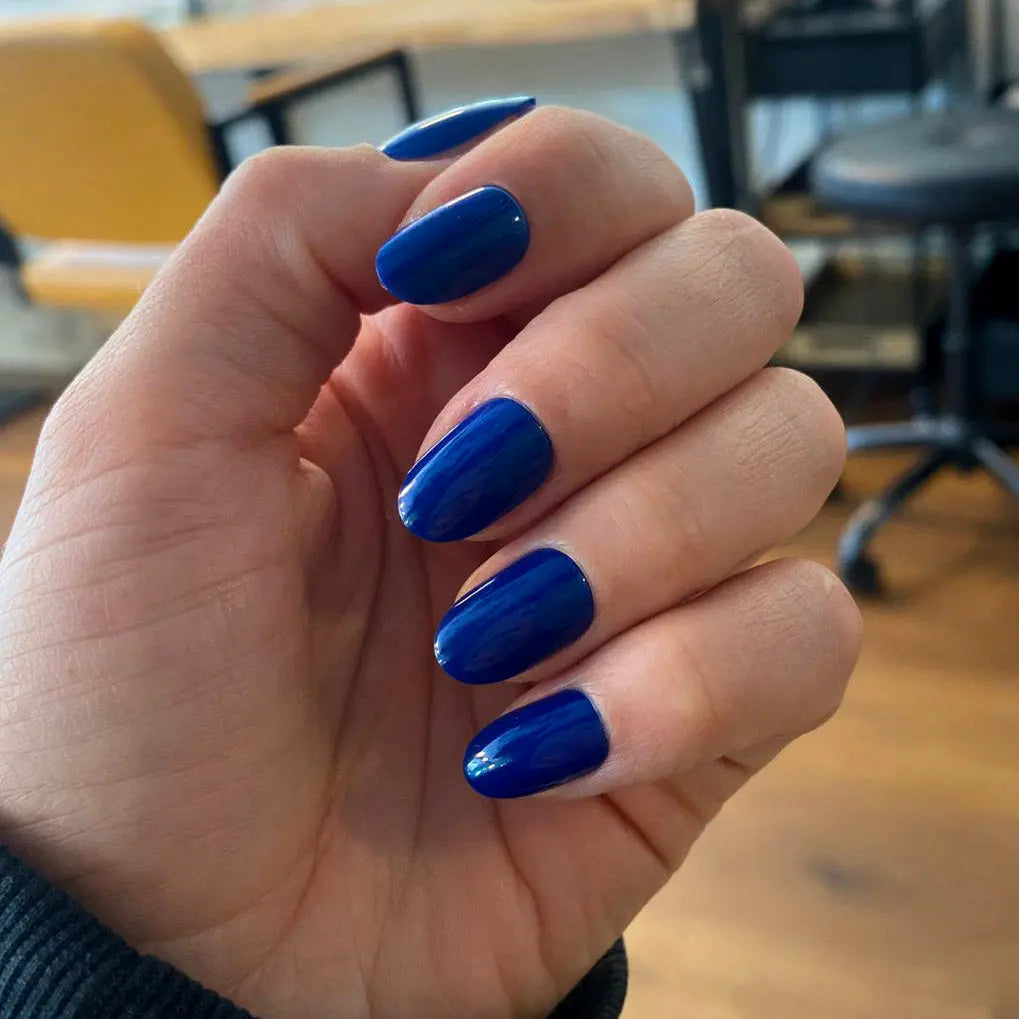 Press-on Nails Bleu Royal arrondis courts sur main au bureau, chaises colorées en fond, faux ongles bleu cobalt vifs et affirmés LYLY Nails