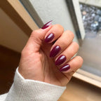 Press-on Nails Bordeaux Velours amande courts sur main près d'une fenêtre, pull blanc en maille, teinte bordeaux profonde et sophistiquée LYLY Nails