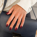 aux ongles Bordeaux Velours amande courts sur main posée sur un sac cuir bleu marine, bague fine dorée et pull torsadé blanc, manucure bordeaux chic et hivernale LYLY Nails