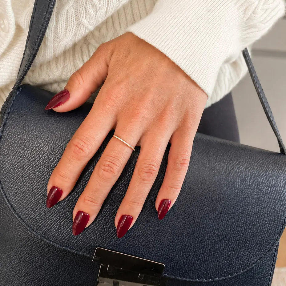 aux ongles Bordeaux Velours amande courts sur main posée sur un sac cuir bleu marine, bague fine dorée et pull torsadé blanc, manucure bordeaux chic et hivernale LYLY Nails