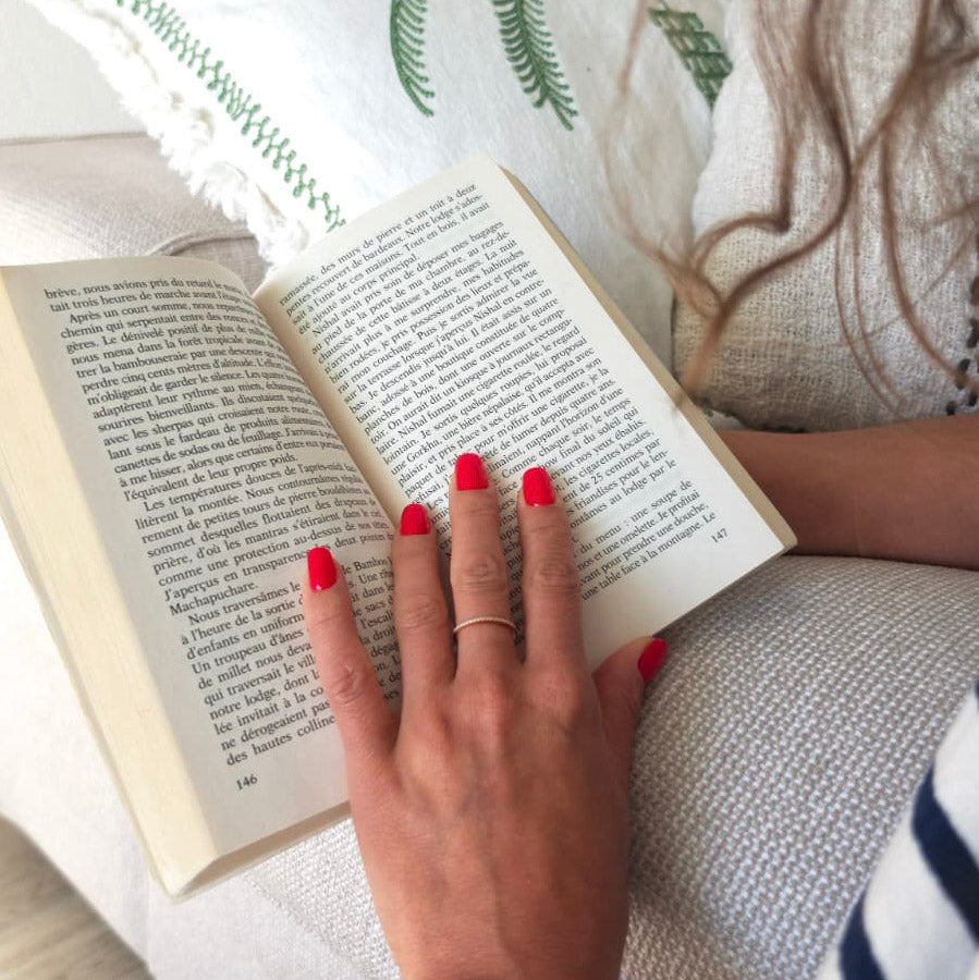 Faux ongles Corail Rosé carré court sur main posée sur un livre, fond lin blanc et motifs verts, manucure rouge corail lumineuse et féminine LYLY Nails