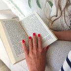 Faux ongles Corail Rosé carré court sur main posée sur un livre, fond lin blanc et motifs verts, manucure rouge corail lumineuse et féminine LYLY Nails