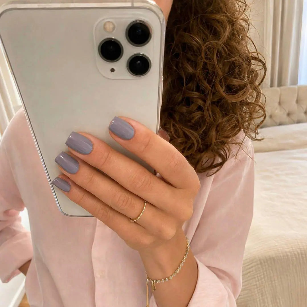 Faux ongles Lavande Nuit carrés courts sur main tenant un iPhone, chemise rose et bracelet doré, cheveux bouclés, ambiance chambre cosy, manucure gris-mauve sophistiquée LYLY Nails