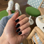 Faux ongles Noir Intense forme carrée longueur courte sur main décontractée en café, tabourets verts et rotin, style urbain chic LYLY Nails