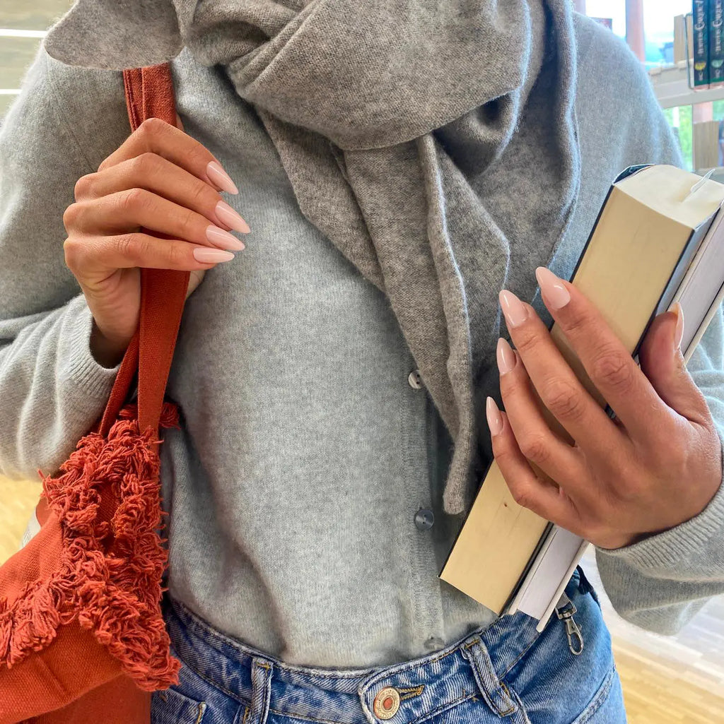 Manucure Press-on Pêche Douce amande moyen sur deux mains en bibliothèque, sac terracotta à franges et livre, gilet et écharpe gris, ambiance cosy automne LYLY Nails