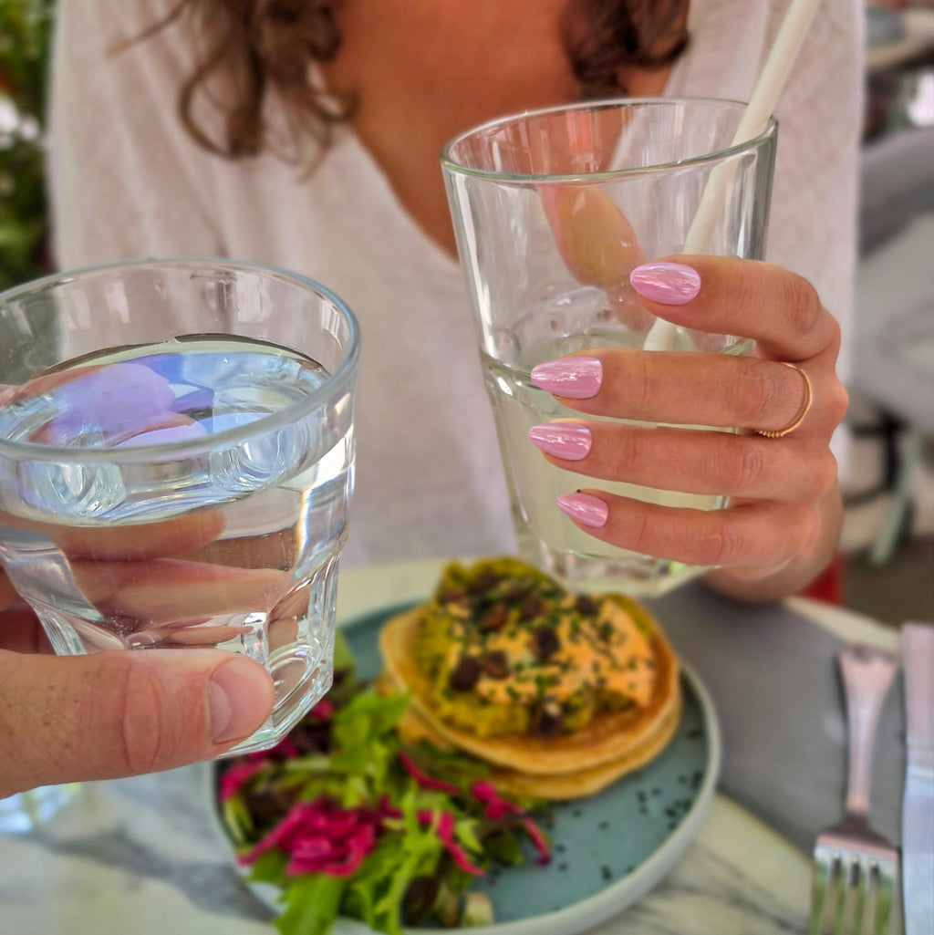 Press-on Nails Rose Orchidée carrés amande courts, main tenant un verre d'eau en terrasse brunch, assiette colorée en fond, faux ongles rose vif et joyeux LYLY Nails