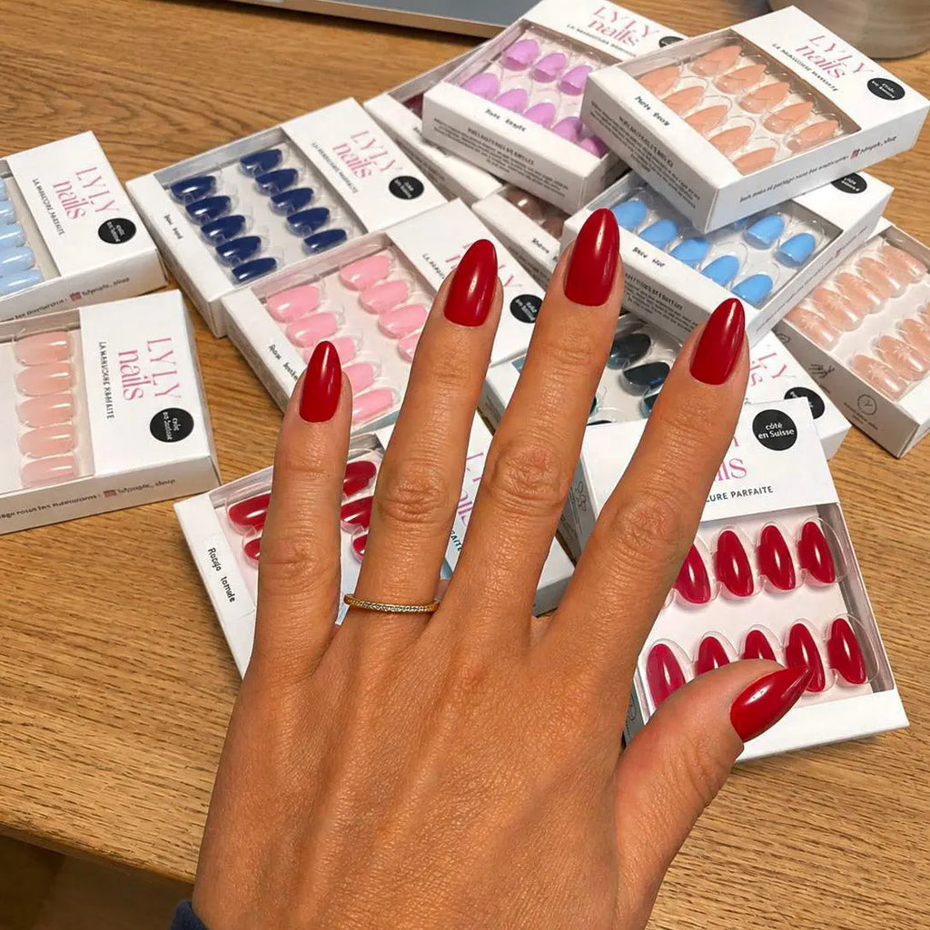 Manucure Press-on Rouge Coquelicot amande courte, main posée sur une table entourée de boîtes LYLY Nails, toute la collection visible, ongles rouges vifs et féminins LYLY Nails