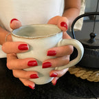 Faux ongles Rouge Framboise carrés courts, deux mains autour d'une tasse en céramique, théière fonte noire et ambiance cuisine chaleureuse, ongles rouges intenses LYLY Nails