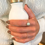 Press-on Nails Taupe Chic carrés courts sur main tenant une gourde blanche, pull torsadé blanc et deux bagues dont une pierre verte, manucure taupe naturelle et élégante LYLY Nails